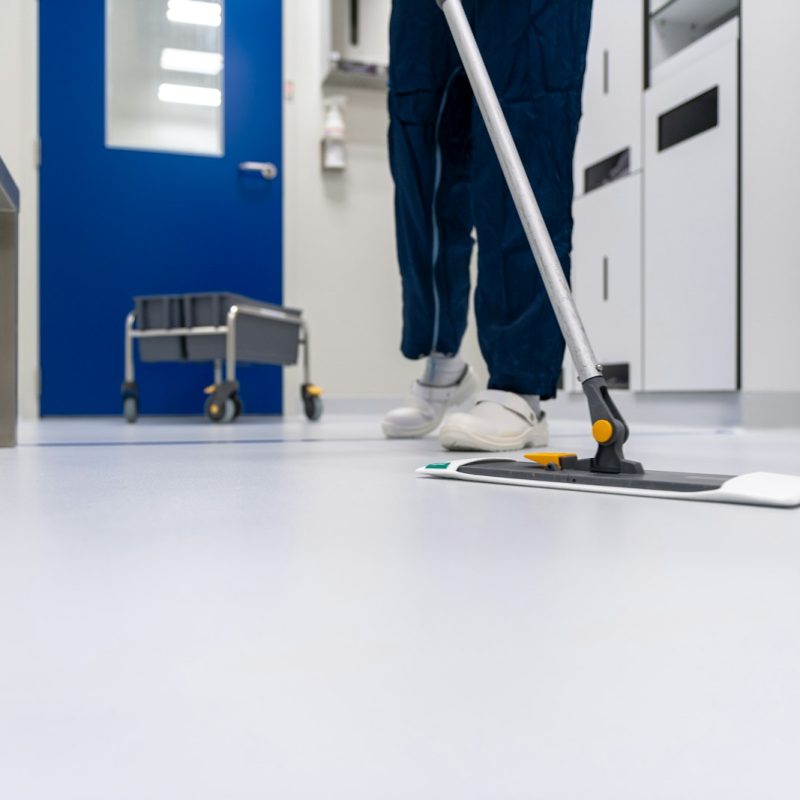 Flat microfibre mop being pushed across a pale grey hard floor, with a trolley and blue door in the background