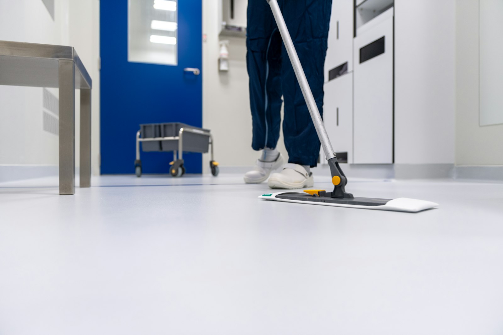 Flat microfibre mop being pushed across a pale grey hard floor, with a trolley and blue door in the background